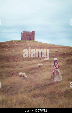 Una ragazza in un abito vintage su un prato con le pecore, sullo sfondo di una cappella Foto Stock