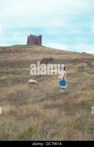 Una ragazza in un abito vintage su un prato con le pecore, sullo sfondo di una cappella Foto Stock