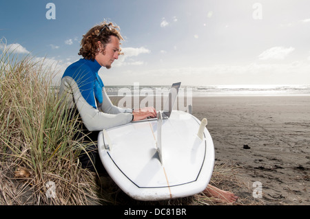 Surfista maschile seduto sulla spiaggia con il computer portatile sulla tavola da surf, Ngarunui Beach, Raglan, Nuova Zelanda Foto Stock