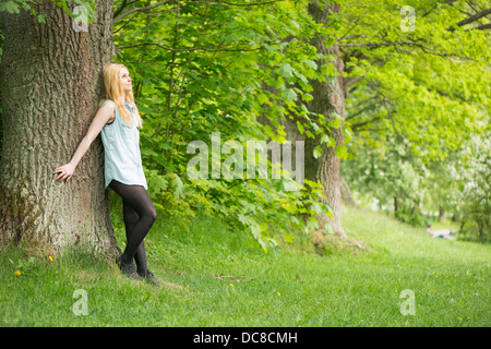 La natura in scena con una giovane donna attraente appoggiata contro un albero in un parco Foto Stock