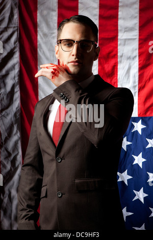 Un uomo vestito come un banchiere con un volto insanguinato in piedi di fronte a una bandiera americana, appeso a testa in giù, poste dal modello. Foto Stock