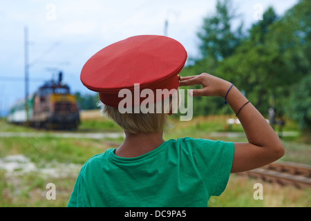 Bambino ragazzo biondo con Train Dispatcher cappuccio della riproduzione nella stazione ferroviaria Foto Stock