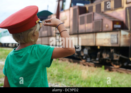 Bambino ragazzo biondo con Train Dispatcher cappuccio della riproduzione nella stazione ferroviaria Foto Stock