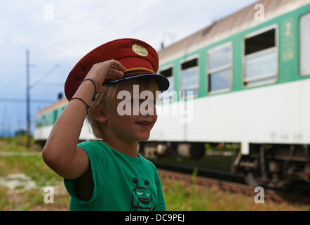 Bambino ragazzo biondo con Train Dispatcher cappuccio della riproduzione nella stazione ferroviaria Foto Stock