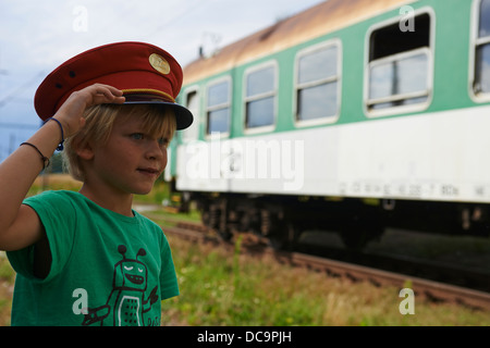 Bambino ragazzo biondo con Train Dispatcher cappuccio della riproduzione nella stazione ferroviaria Foto Stock