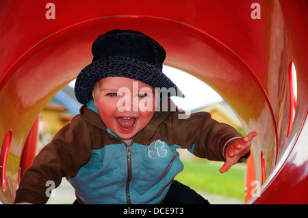 Un sorridente baby boy (di età compresa tra i 16 mesi) in un rosso del tubo di plastica in un parco giochi Foto Stock