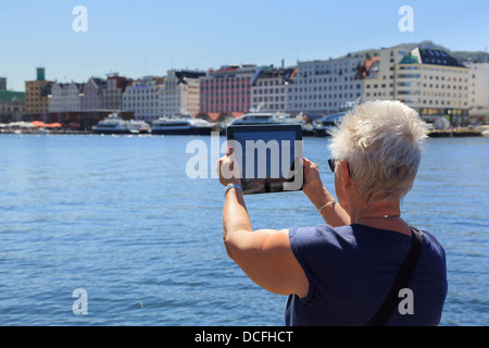 Donna senior turista baby-boomer generazione tenere un iPad tablet a lunghezza d'armi per scattare una fotografia del porto di Vagen Bergen Hordaland Norvegia Foto Stock