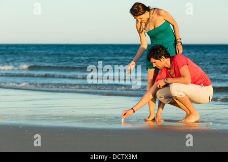 L uomo e la donna, giovane, godendo del romantico tramonto su una spiaggia dall'oceano nella loro vacanza, essi gusci di ricerca Foto Stock