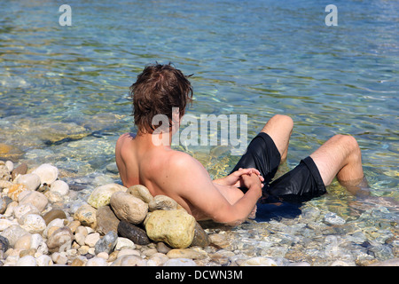 19 anno vecchio ragazzo puntellato e rilassante contro una pietra torna poggiare su una spiaggia di ciottoli in Grecia Foto Stock