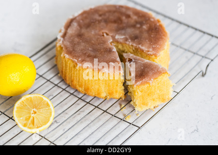 Home limone al forno cospargere la torta con la glassa di zucchero superiore, con una fetta tagliata su una griglia di raffreddamento con due limoni Foto Stock