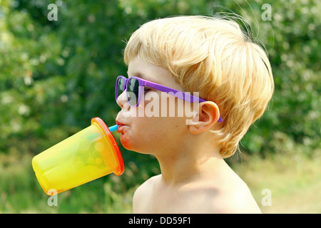 Un giovane ragazzo che indossa gli occhiali da sole viola si inclina indietro la sua testa bere il succo da un colorato sippy cup su una soleggiata giornata estiva Foto Stock