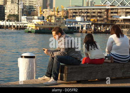 Un giovane uomo seduto su di un molo harbourside, guardando un mobile (cellulare) telefono, Darling Harbour, Sydney, Australia Foto Stock