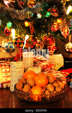 Ciotola di vetro riempito con le noci, le nocciole e mandarini arance con i regali di Natale sotto la struttura ad albero per la parte posteriore. Foto Stock