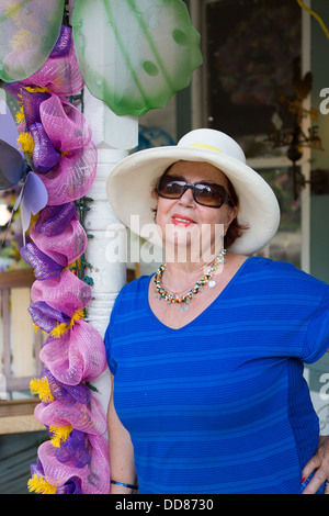 Elegante donna senior in occhiali da sole e una vasta colmato cappello di paglia appoggiata contro un palo a fianco di decorativi fiori rosa Foto Stock