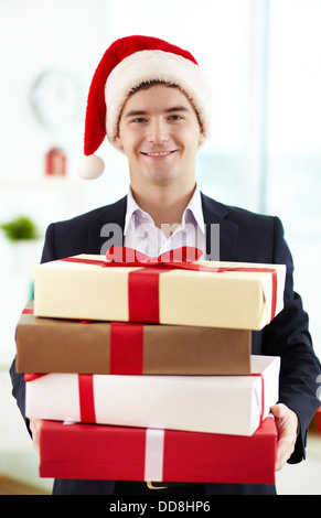 Immagine di happy businessman in Santa trattenimento tappo pila di doni e guardando la telecamera in ufficio Foto Stock