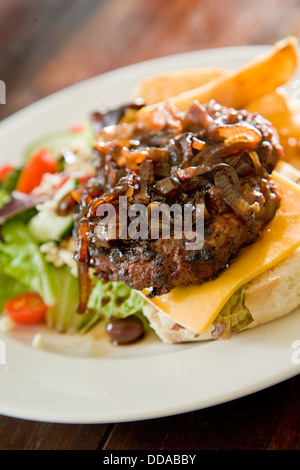 Grigliate di carni bovine patty, guarnita con le cipolle caramellate, sulla fetta di ricca di formaggio cheddar, con fresca insalata greca, frittura di patatine fritte. Foto Stock