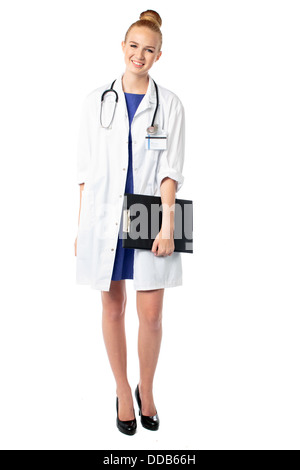 Sorridente attraente giovane medico donna in piedi tenendo un file di record paziente nella sua mano come lo fa la sua ward rounds, isolato su bianco Foto Stock