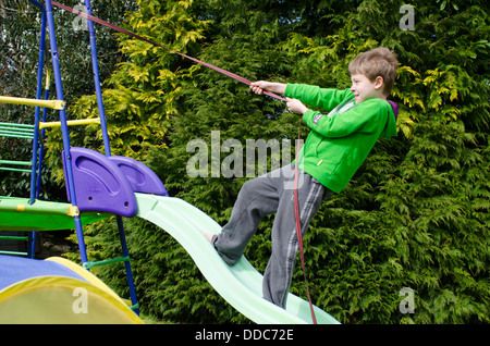 Bambino di otto anni salendo su un giardino di contenimento scorrevole di una fune o cinghia che egli ha vincolato al telaio Foto Stock