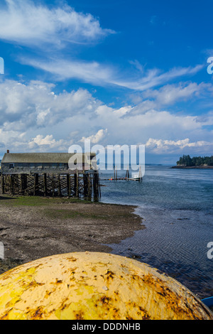 Una vista del Lubec, Maine seashore con un grande giallo del galleggiante in metallo in primo piano e un vecchio impianto di pesce in background. Foto Stock