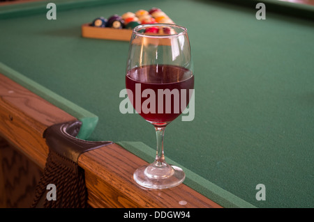 Vino rosso in un singolo in vetro trasparente su un tavolo da biliardo con un rack della piscina di palline in background seduto su un tavolo in legno di quercia feltro verde. Foto Stock