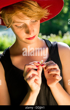 Giovane donna, che indossa un feltro di Red Hat e un abito nero, tirando delicatamente verso i petali di una margherita Foto Stock