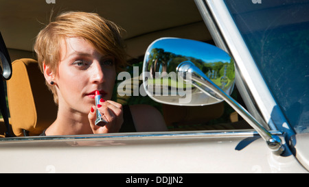 Giovane donna mettendo il rossetto in lo specchio retrovisore di un classico auto oldtimer Foto Stock