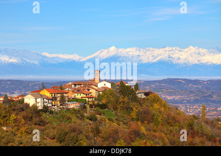 Vista della piccola cittadina sulla cima di una collina circondata da alberi autunnali e vette innevate delle Alpi su sfondo in Piemonte, Italia. Foto Stock