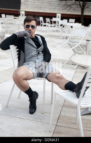 Cheeky giovane uomo nel quartiere alla moda di occhiali che indossa pantaloncini grigio con gambe pelose seduti su una sedia a street cafe Foto Stock