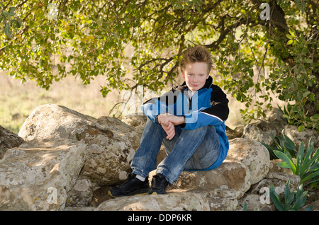 Ragazzo seduto in natura su una roccia sotto un albero Foto Stock