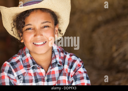 Bella e felice razza mista americano africano ragazza femmina bambino paglia che indossa il cappello da cowboy & plaid shirt seduta nel fieno riempito barn Foto Stock