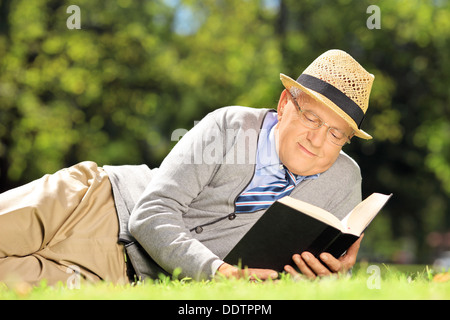 Senior uomo con hat sdraiati su un prato e la lettura di un libro in un parco Foto Stock