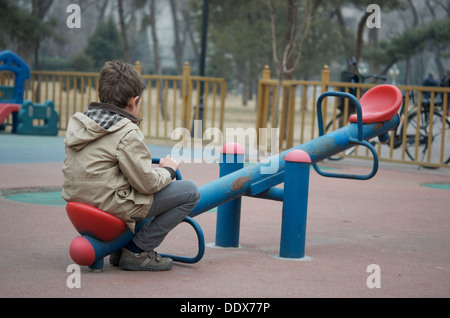 Un piccolo ragazzo seduto da solo su un seasaw presso un parco giochi vuoto