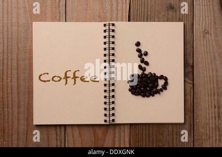 Un gruppo di chicchi di caffè conformato in una tazza di caffè sulla pagina vuota di un notebook. La pagina di fronte ha la parola caffè scritto Foto Stock