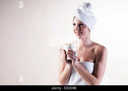 Sorridente giovane donna con un asciugamano avvolto attorno alla sua testa tenendo una tazzina Foto Stock