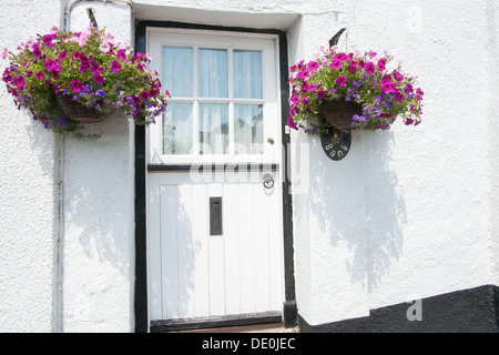 Porta bianca con telaio nero, parete dipinta di bianco e cesti floreali pendenti in una stretta viuzza in Dunsford, REGNO UNITO Foto Stock