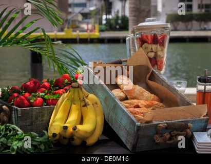 Impostazione urbana, prima colazione dall'acqua. Frullatore con un trito di frutta sulla tavola con scatola di beni cotti al forno, fragole e banane. Foto Stock