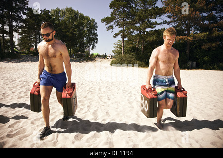Due dure giovane maschio crossfitters sollevamento i bidoni di pesanti e camminare durante un allenamento crossfit sulla spiaggia. Foto Stock