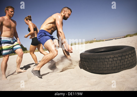 Forte atleta maschio circa per capovolgere un carrello pneumatico. I giovani facendo esercizio crossfit sulla spiaggia in una giornata di sole. Foto Stock