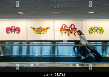 Mostra Art of Recology Aeroporto di San Francisco | SAN FRANCISCO, California - quattro maschere a parete dell'artista Barbara Holmes sono appese sopra un passaggio pedonale mobile nel Terminal 3 dell'Aeroporto Internazionale di San Francisco, realizzate con materiali recuperati attraverso il programma Artist in Residence di Recology. Le maschere fanno parte del "Art of Recology: The Artist in Residence Program 1990-2013", una mostra costruita interamente a partire dal flusso di rifiuti scartati dalla città. Un viaggiatore passa sotto tirando i bagagli. Foto Stock