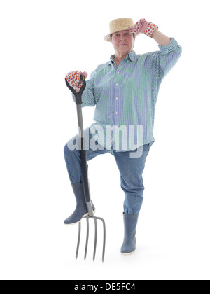 Senior giardiniere donna indossa cappello di paglia e stivali di gomma in posa con forche su sfondo bianco Foto Stock