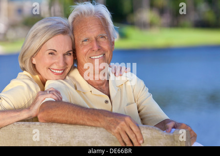 Sorridenti romantico senior l uomo e la donna giovane seduto su una panchina nel parco che abbraccia da un lago blu Foto Stock