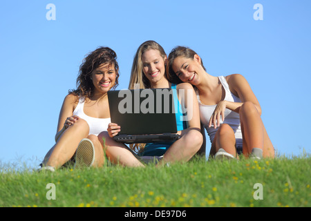 Un gruppo di tre ragazze teenager di ridere mentre guardi il laptop seduta sul prato con il cielo in background Foto Stock