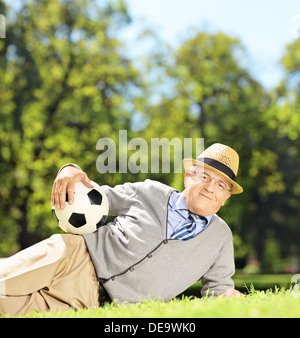 Senior uomo con hat sdraiati su un prato verde e trattenimento di un pallone da calcio in un parco Foto Stock