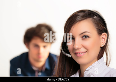 Ritratto di un giovane smart call center dipendente di sesso femminile a sorridere a voi in un ufficio luminoso con il collega di sesso maschile in background. Foto Stock