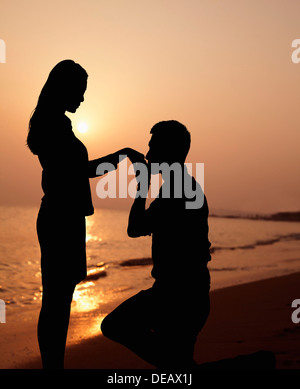 Silhouette di un ragazzo in ginocchio e baciare la sua mano fidanzate sulla spiaggia al tramonto Foto Stock