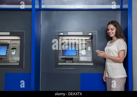 Sorridente giovane donna in piedi di fronte a un bancomat e guardando il suo cellulare Foto Stock