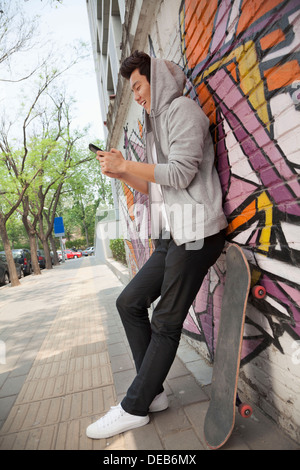 Giovane ancora pendente un muro coperto di graffiti e messaggi di testo sul suo telefono, skateboard è accanto a lui Foto Stock