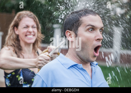 Giovane giocando con un tubo flessibile da giardino e la spruzzatura di ogni altro fuori in giardino, l'uomo ha un sguardo scioccato, close-up Foto Stock