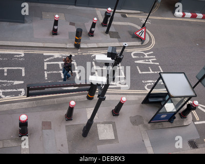 Una donna attraversa le barriere stradali e le telecamere a circuito chiuso di sorveglianza nella City di Londra, Regno Unito, evidenziando la presenza di infrastrutture di sicurezza nel distretto finanziario della capitale. Foto Stock