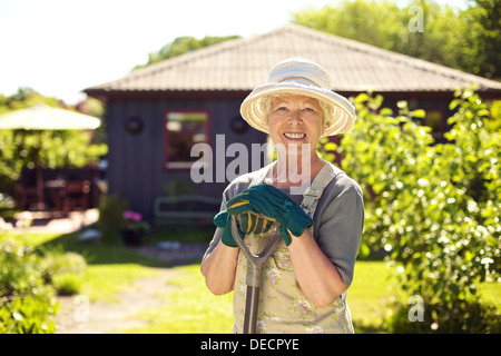 Ritratto di Allegro senior donna con utensili da giardinaggio all'esterno. Donna anziana in piedi con la pala nel suo giardino nel cortile Foto Stock